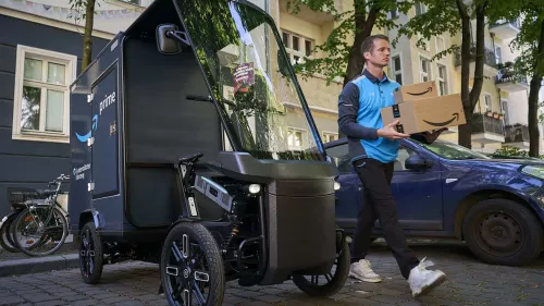 Amazon-Lieferant in der Berliner Innenstadt auf einem Parkplatz mit Cargo PACK und zwei Paketen in der Hand.